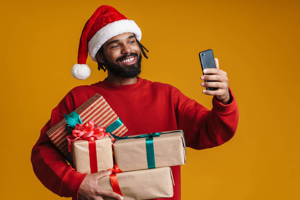 Happy african american guy taking selfie on cellphone and holding Christmas gifts isolated over yellow background - Фото, изображение
