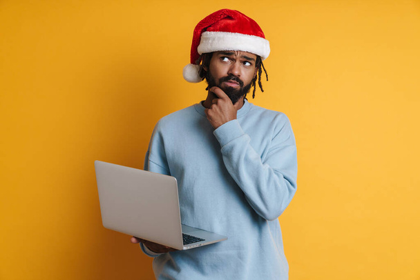 Pensive mladý santa africký Američan muž v vánoční klobouk pracuje na notebook PC izolované přes žluté pozadí - Fotografie, Obrázek