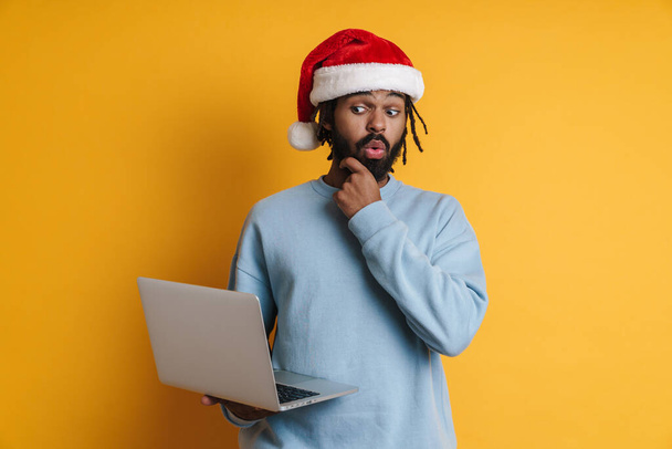 若いですサンタアフリカ系アメリカ人男性でクリスマスの帽子作業ノートパソコンPCコンピュータ上で隔離された黄色の背景 - 写真・画像