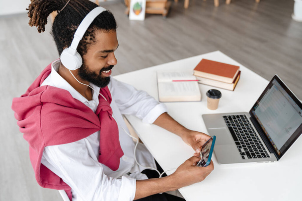 Cheerful african american guy in headphone using cellphone and working with laptop in office - Photo, Image