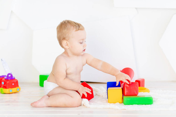 malé dítě chlapec na šest měsíců hraje s jasnými kostkami v jasném bílém pokoji v plenkách - Fotografie, Obrázek