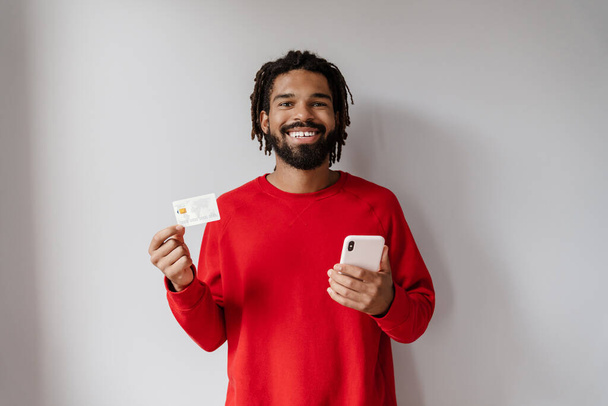 Joyful african american man posing with cellphone and credit card isolated over white background - Valokuva, kuva