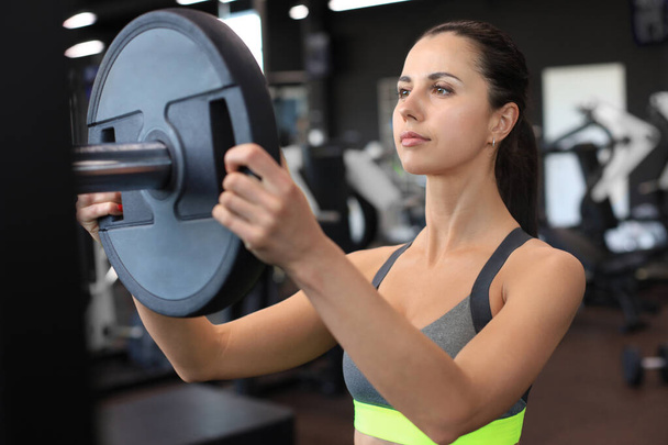 Strong young woman with beautiful athletic body adding weight on barbell at gym - Foto, Imagem