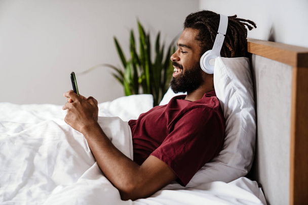 Cheerful african american man in headphones using cellphone while lying in bed at home - 写真・画像