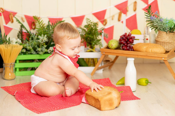 Ένα μικρό παιδί. Ένα αγόρι έξι μηνών κάθεται στο πάτωμα της κουζίνας με ένα καρβέλι ψωμί. - Φωτογραφία, εικόνα