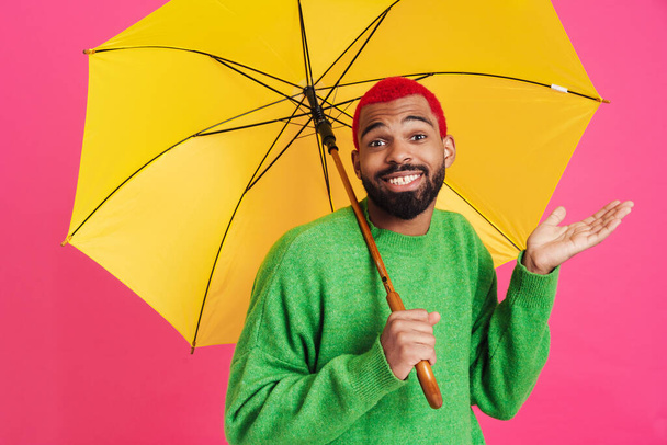 Joyful african american guy holding copyspace while posing with umbrella isolated over pink background - Foto, afbeelding