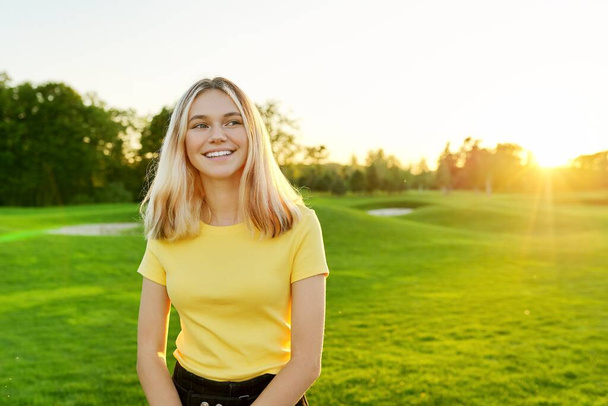 Outdoor portrait of smiling teenage girl 16, 17 years old in yellow T-shirt, on green sunny lawn - Valokuva, kuva