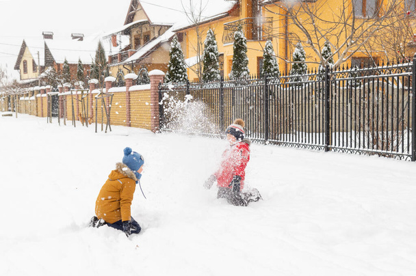 Dwóch chłopców ubranych w żółte i czerwone kurtki zimowe bawiących się na zewnątrz w śnieżki. Zimowy zimny dzień, wszystko pokryte jest śniegiem, ponura pogoda - Zdjęcie, obraz