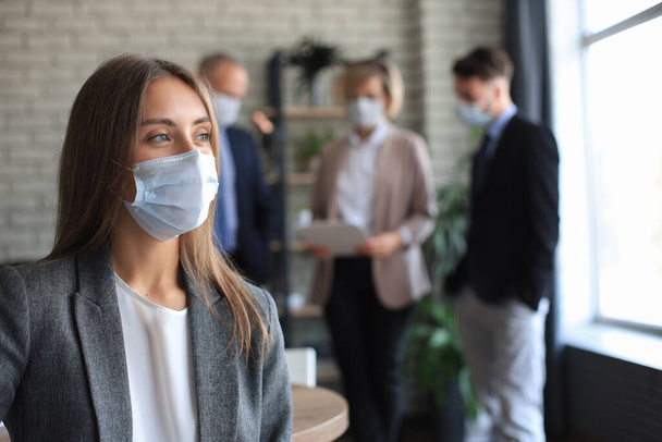 Imprenditrice in maschera medica con il suo personale, gruppo di persone in background in un moderno ufficio luminoso all'interno - Foto, immagini