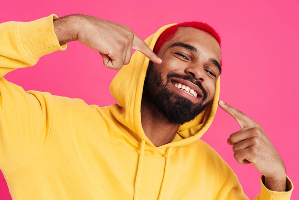 Joyful african american guy smiling and pointing fingers at his cheeks isolated over pink background - Фото, изображение