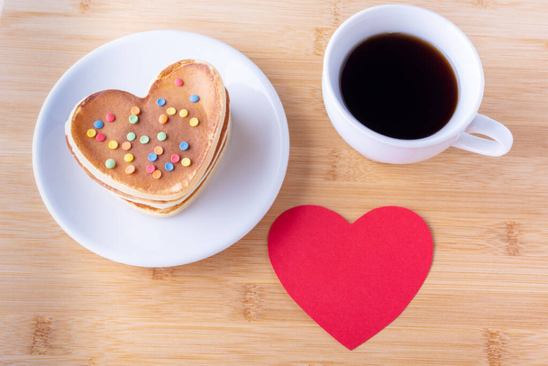 Creative breakfast for Valentines Day. Homemade heart shaped pancakes with sugar decoration on white plate, fork, mug with coffee or cocoa and red empty heart shape - Photo, Image