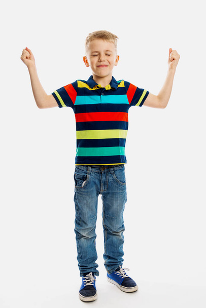 A blond boy in jeans and a multicolored T-shirt stands with his eyes closed and his hands up. White background. Vertical - 写真・画像