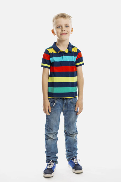 A blond boy in jeans and a multicolored T-shirt stands. White background. Vertical. Full height. - Fotografie, Obrázek