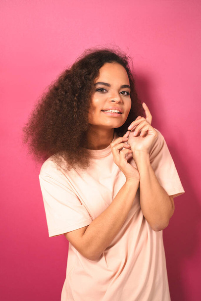 Güzel koyu tenli Afro-Amerikalı genç kız pembe arka planda izole edilmiş şeftali renkli tişört giymiş kameraya nazikçe bakıyor. Güzellik konsepti. Yüz ifadeleri, duygular, duygular - Fotoğraf, Görsel