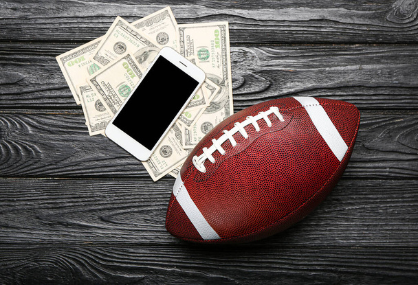 Dinero, teléfono móvil y pelota de rugby sobre fondo de madera oscura. Concepto de apuesta deportiva - Foto, imagen