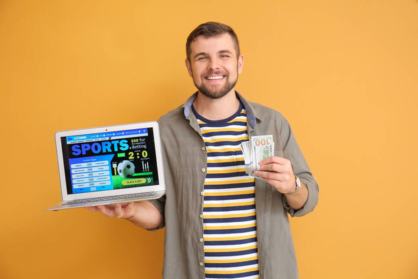Young man with money and laptop placing sports bet on color background - Photo, Image