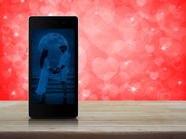 Romantic loving couple standing and holding hands together on modern smart mobile phone screen on wooden table over blur red love heart background, Business internet dating online, Valentines day concept - Foto, Imagem