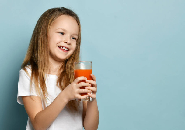 Cheerful happy smiling cute kid girl in white t-shirt standing with glass of fresh fruit juice and looking at copy space - Fotoğraf, Görsel