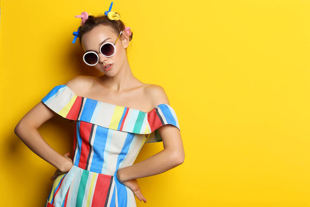 Fashion cool girl posing in sunglasses on yellow background. Young hipster woman, curlers in her hair  - 写真・画像