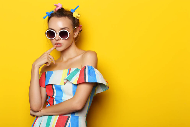 Fashion cool girl posing in sunglasses on yellow background. Young hipster woman, curlers in her hair  - Фото, зображення