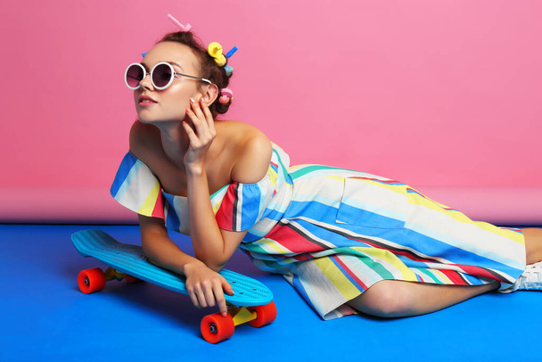 Fashion cool girl posing with skateboard. Young hipster woman, curlers in her hair, sunglasses, white sneakers. studio shot  - 写真・画像
