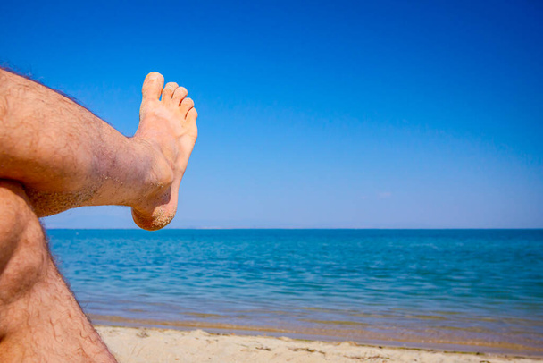Les jambes croisées de l'homme bronzent en s'allongeant sans soucis sur le sable à côté du littoral, sur la plage publique. - Photo, image