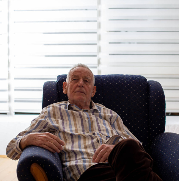 Homme âgé assis dans un fauteuil et regardant fixement dans un point à la maison. Concept de solitude et de démence - Photo, image