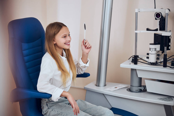 Jeune femme optimiste en vêtements décontractés assis dans une chaise de médecine bleue tout en regardant vers l'avenir dans le centre de médecine - Photo, image