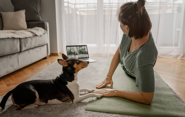 Cheerful beautiful woman practicing yoga at home online on the carpet and yoga mate with her dog nearby, sports and mental health - Valokuva, kuva