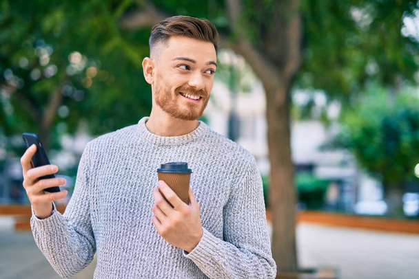 Jonge blanke man met smartphone die koffie drinkt in het park. - Foto, afbeelding