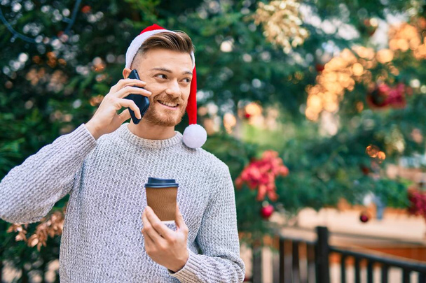 公園でクリスマスの帽子をかぶった若い白人男性がスマートフォンで話してコーヒーを飲んでいます - 写真・画像