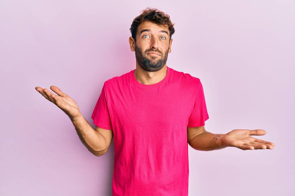 ピンクの背景にカジュアルなピンクのTシャツを着た髭を生やしたハンサムな男は、腕や手で上げられた混乱した表情。疑わしい概念.  - 写真・画像