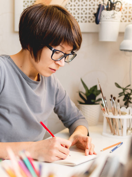 Woman with short hair cut is drawing in notebook. Calming hobby, anti stress leisure. Artist at work. Cozy workplace. - Photo, Image