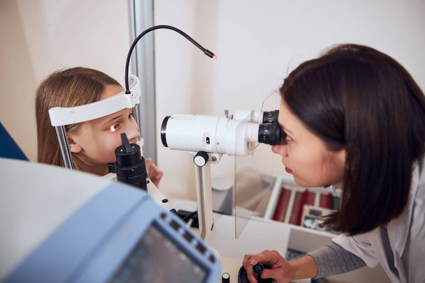 Adolescente loira olhando para o médico durante a verificação de sua visão com oftalmoscópio - Foto, Imagem
