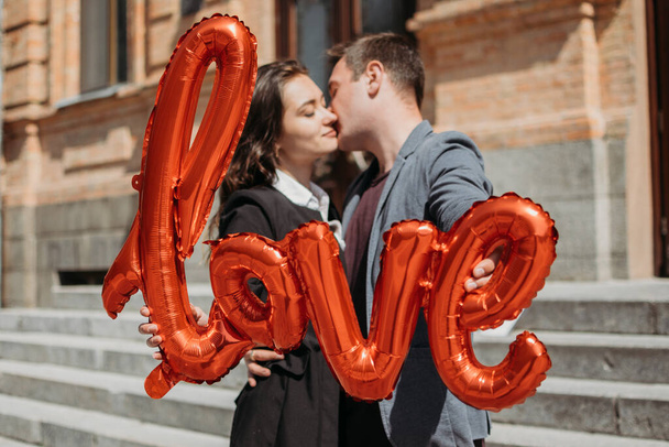 Walentynki świętowanie i randki koncepcja. Szczęśliwa kochająca się para z czerwonymi balonami miłości na ulicy miasta. Odkryty portret młodej pary z balonem słowa Miłość. - Zdjęcie, obraz