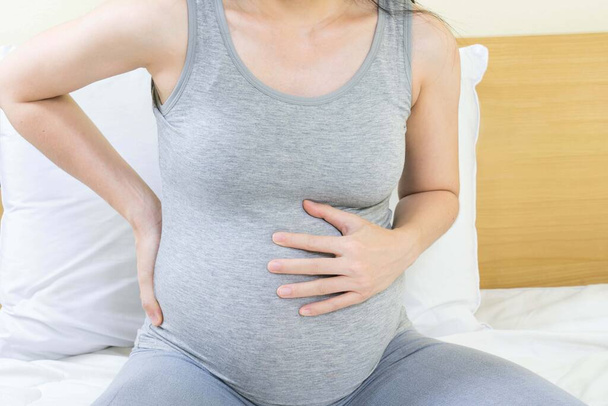Feeling bad concept - Close up of pregnant woman with back pain and stomachache sitting on bed at home - Фото, зображення