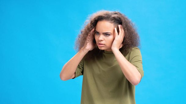 青い背景に隔離されたオリーブのTシャツを着て頭痛に苦しんでいる彼女の頭に触れるストレスや不幸なアフリカ系アメリカ人の少女。美しさの概念。ヘルスケアの概念 - 写真・画像