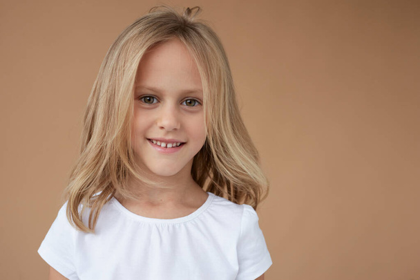 Closeup front portrait of pretty little girl with wavy blond hair, dressed in white shirt, over beige background. - 写真・画像