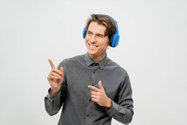 Happy smiling young guy in grey shirt standing listening music wearing blue wireless headphones pointing fingers sideways. Funny young guy listen to his favourite track or song - Foto, afbeelding