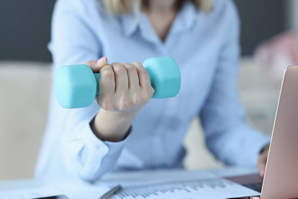 Woman sits on laptop and holds blue dumbbell in her hand - Photo, Image