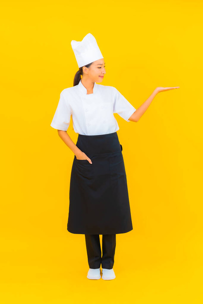 Portret piękna młoda azjatycka kobieta w kucharz lub kucharz mundur z kapeluszem na żółtym odizolowanym tle - Zdjęcie, obraz