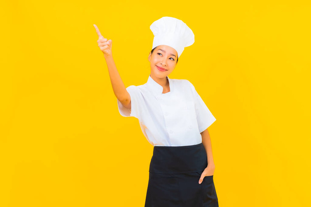 Porträt schöne junge asiatische Frau in Koch-oder Koch-Uniform mit Hut auf gelbem Hintergrund - Foto, Bild