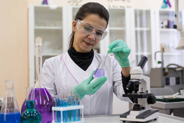 A female scientist in protective glasses and gloves stirs the solution in a flask. Scientific research in a modern laboratory. Creating a vaccine in the laboratory. - Foto, Imagen