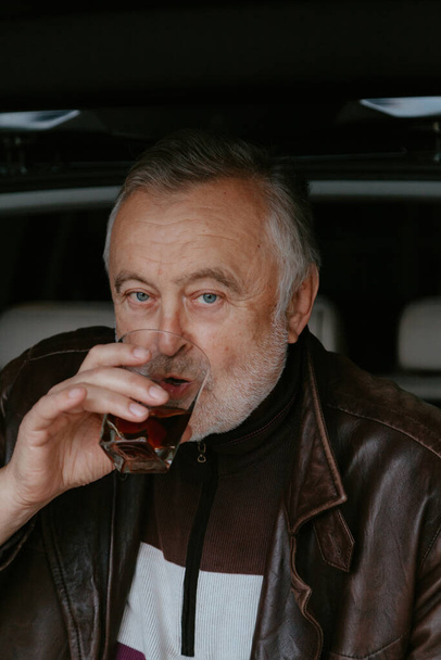 elderly man with a gray beard in a leather jacket drinks whiskey from a glass glass against the background of an open trunk of a passenger ca - Φωτογραφία, εικόνα