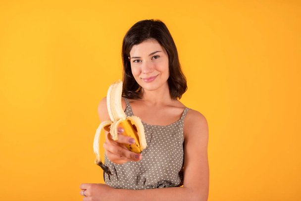 brunette femme en vêtements décontractés manger une banane dans sa main, fond jaune nourriture saine - Photo, image