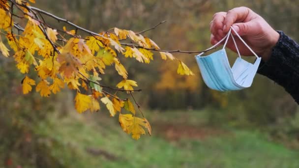 ιατρική μάσκα κρέμεται σε ένα κλαδί ενός φθινοπωρινού δέντρου στο φόντο ενός ανθρώπου. ζωή μετά την καραντίνα. Η ζωή μετά τον κορωναϊό. προστασία από ιούς - Πλάνα, βίντεο