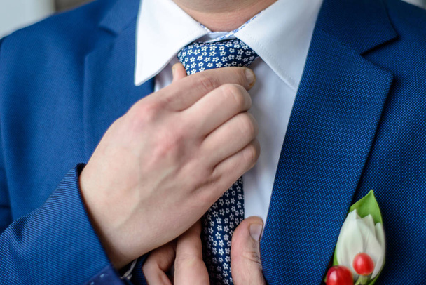 svatební šaty muži: ženich v modrém obleku s boutonniere narovnává kravatu - Fotografie, Obrázek