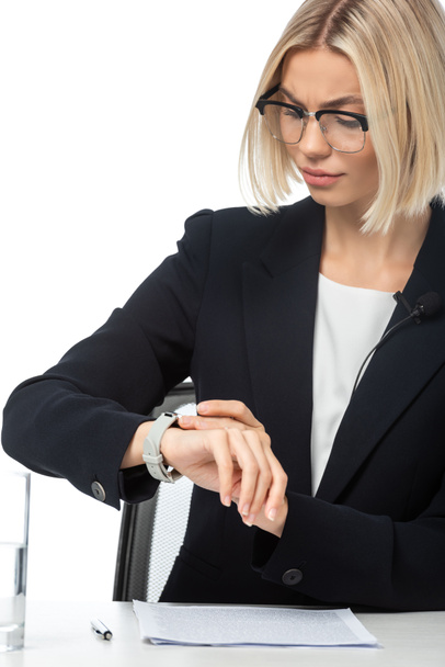 Хмурая ведущая смотрит на наручные часы, сидя на рабочем месте, изолированном на белом - Фото, изображение