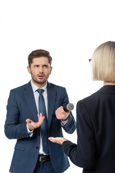 обескураженная журналистка с микрофоном жестикулирует рядом блондинка-бизнесвумен во время интервью изолированы на белом - Фото, изображение
