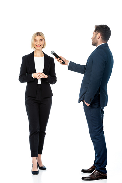 vista completa del presentador con la mano en el bolsillo tomando la entrevista de la mujer de negocios sonriente en blanco - Foto, imagen
